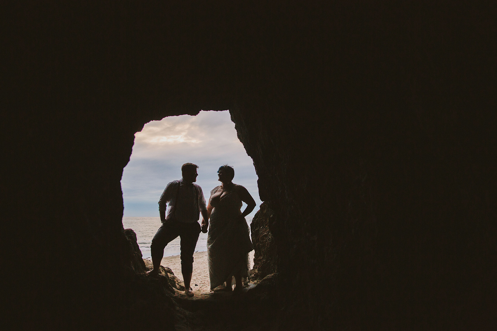Creative portrait of the bride and groom's silhouettes standing in front of the entrance to a cave - Oceanside Community Club Wedding” title=